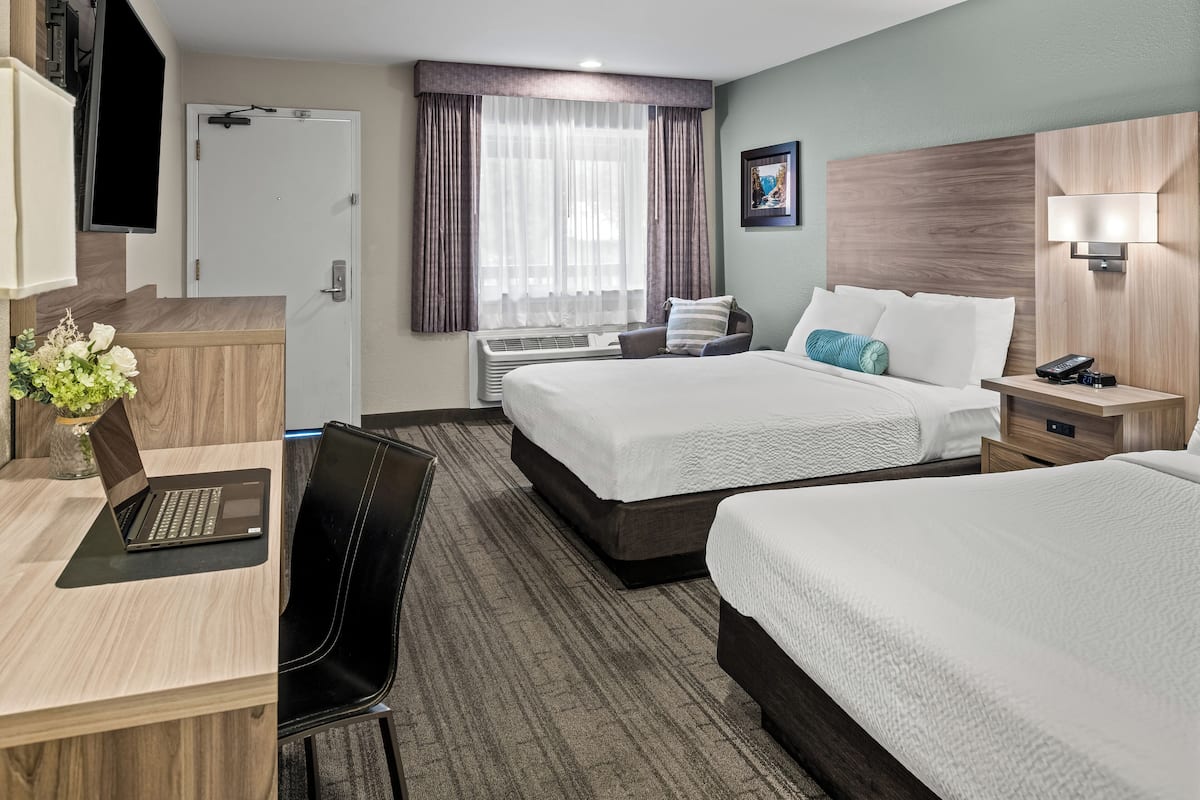 Ein Hotelzimmer mit zwei Betten, einem Schreibtisch mit einem Laptop, einem Stuhl, einem an der Wand montierten Fernseher und einem Fenster mit Vorhängen.