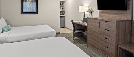 Habitación de hotel con dos camas, un escritorio con computadora, televisor y una kitchenette pequeña.