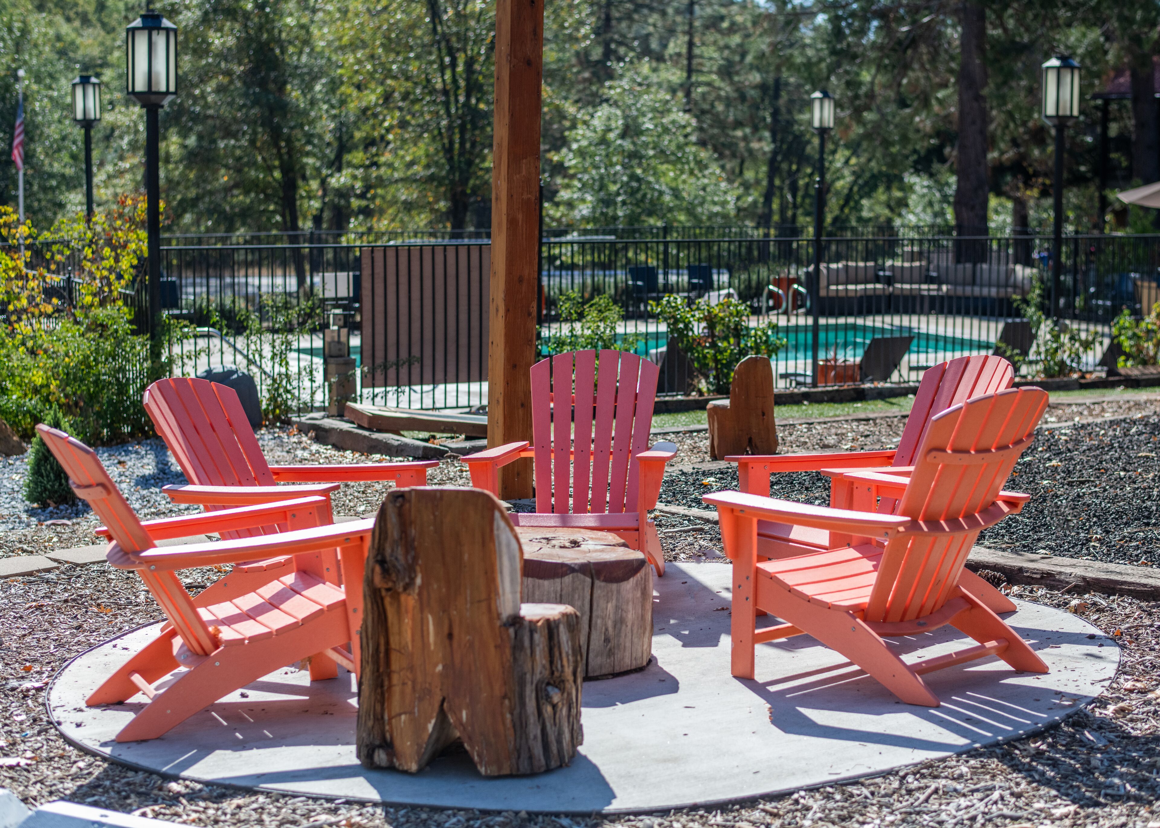 A circular seating area with red Adirondack chairs around a central wooden stump, set on a gravel surface with a pool and greenery in the background.