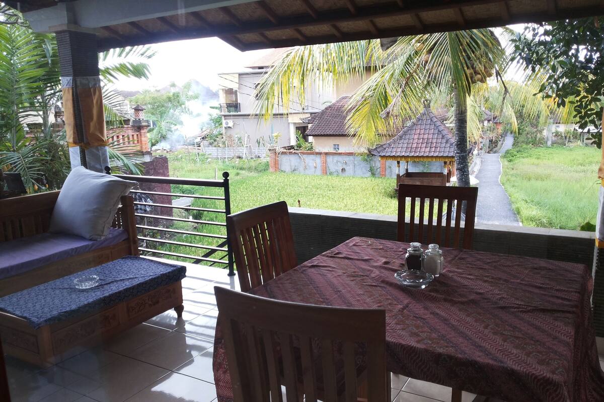 Un espace extérieur abrité, aménagé avec une table et des chaises, un banc rembourré, et offrant une vue sur un jardin et des bâtiments en arrière-plan.