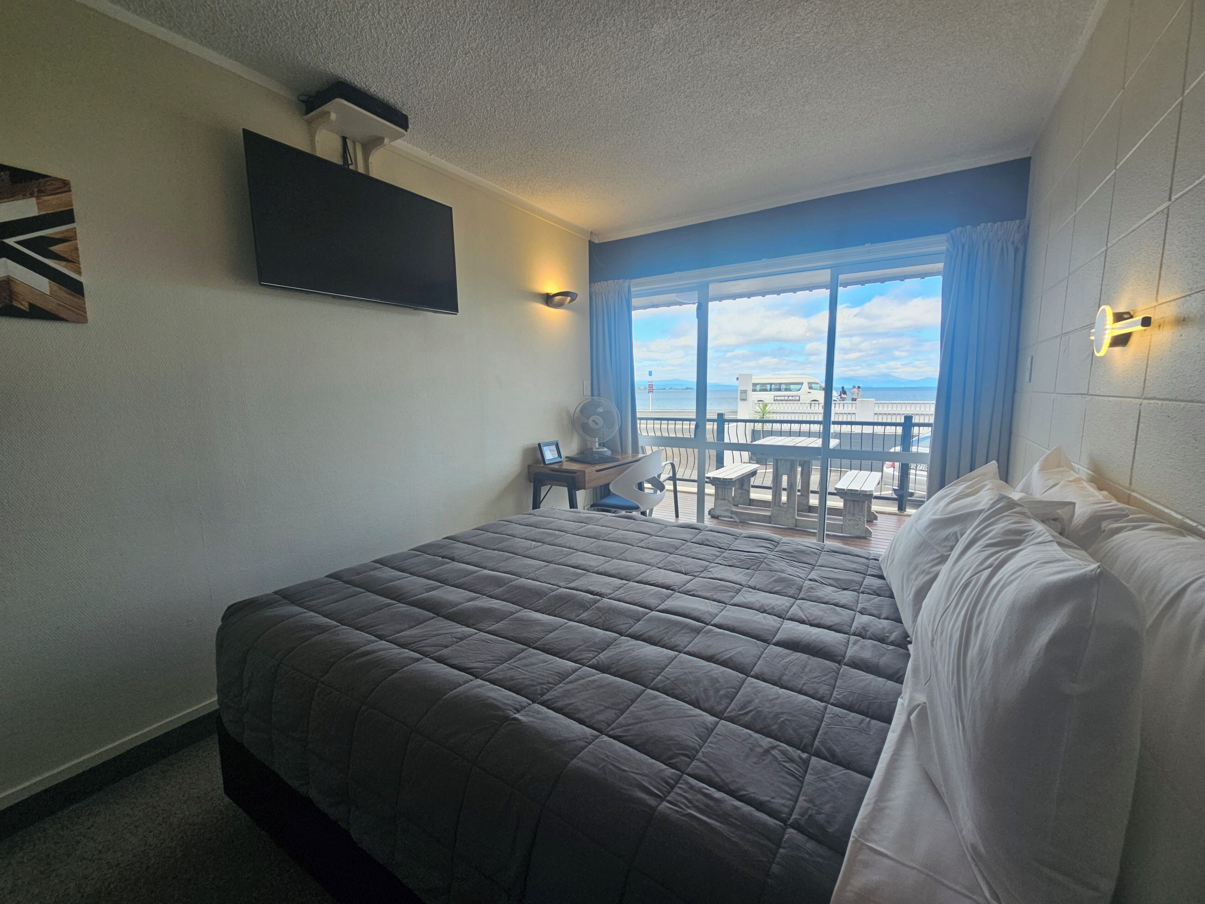 Habitación de hotel con una cama grande, un televisor de pantalla plana en la pared, un escritorio con una silla y un balcón con vista al mar.