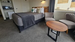 A hotel room with a bed, two armchairs, a small table, a mini-fridge, and a shelf with toiletries.