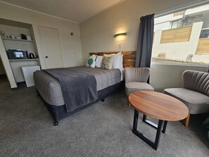 A hotel room with a bed, two armchairs, a small table, a mini-fridge, and a shelf with toiletries. - Tui Oaks Motel (Taupo)