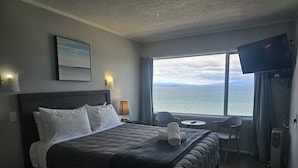 Ein Hotelzimmer mit Bett, Nachttischlampe, Fernseher, Fenster mit Meerblick und einem kleinen Tisch mit Stuhl.