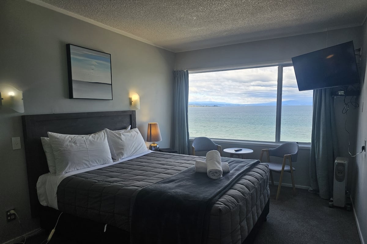 A hotel room with a bed, a bedside lamp, a TV, a window with a view of the sea, and a small table with a chair.