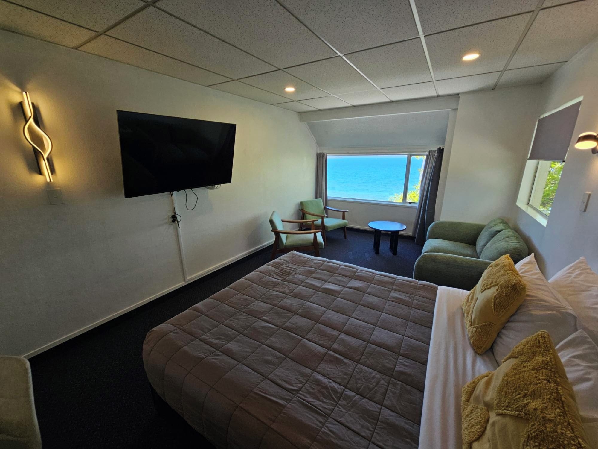 A hotel room with a bed, a TV, a sofa, a chair, and a window with a view of the sea.