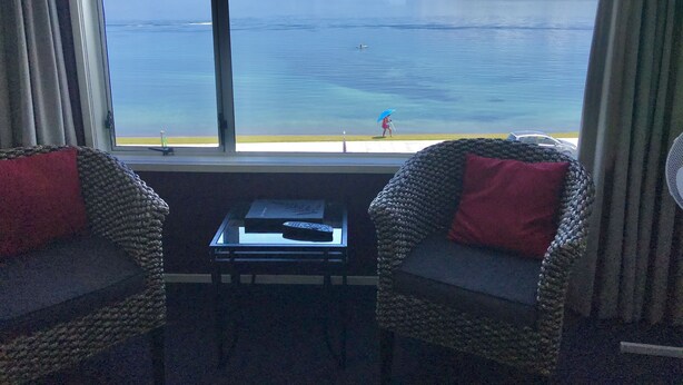Ein Zimmer mit Blick auf einen ruhigen See und Berge, mit zwei Korbstühlen mit roten Kissen, einem kleinen Tisch mit Fernbedienung und einem Fenster mit Vorhängen.