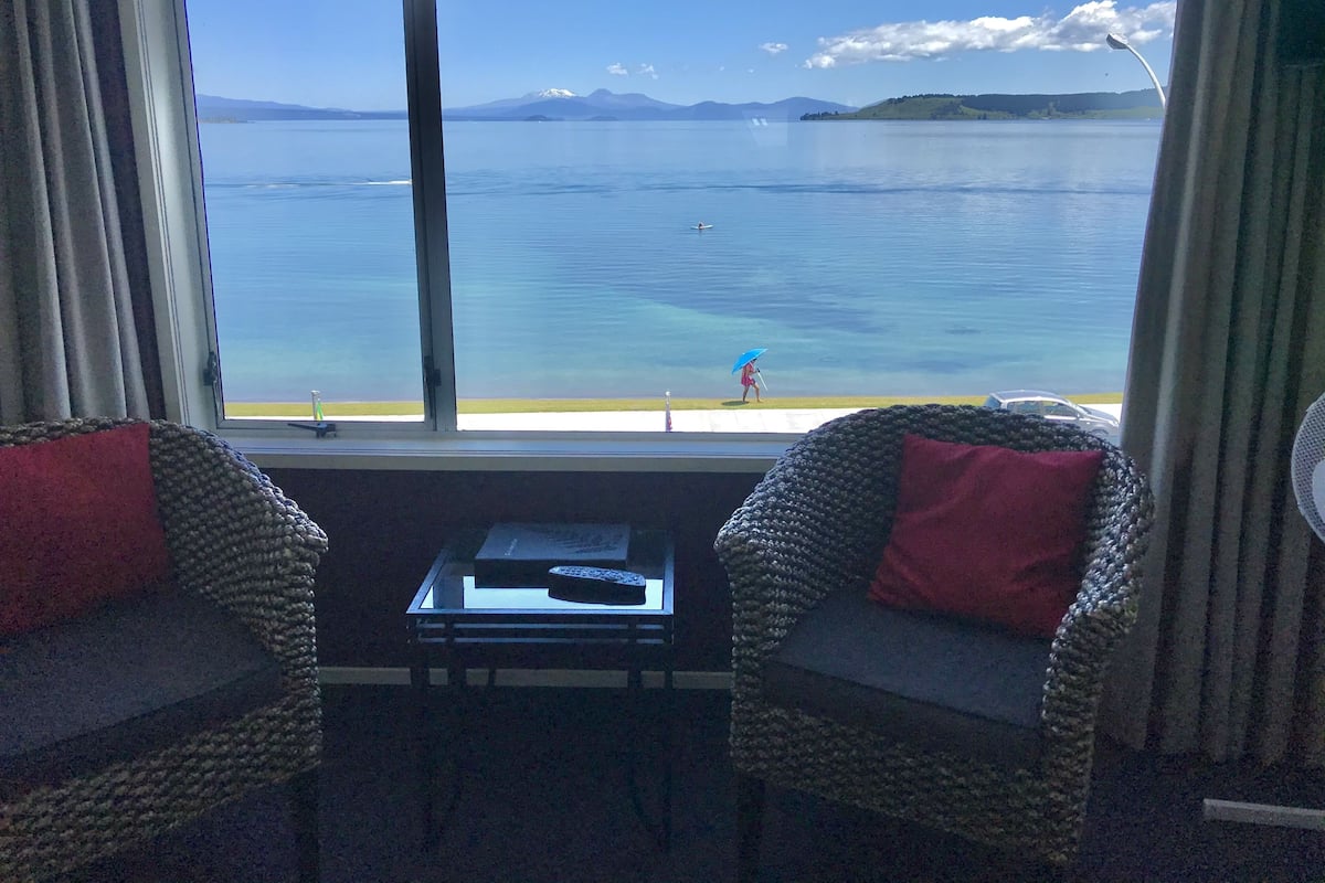 A room with a view of a calm lake and mountains, featuring two wicker chairs with red cushions, a small table with a remote control, and a window with curtains.