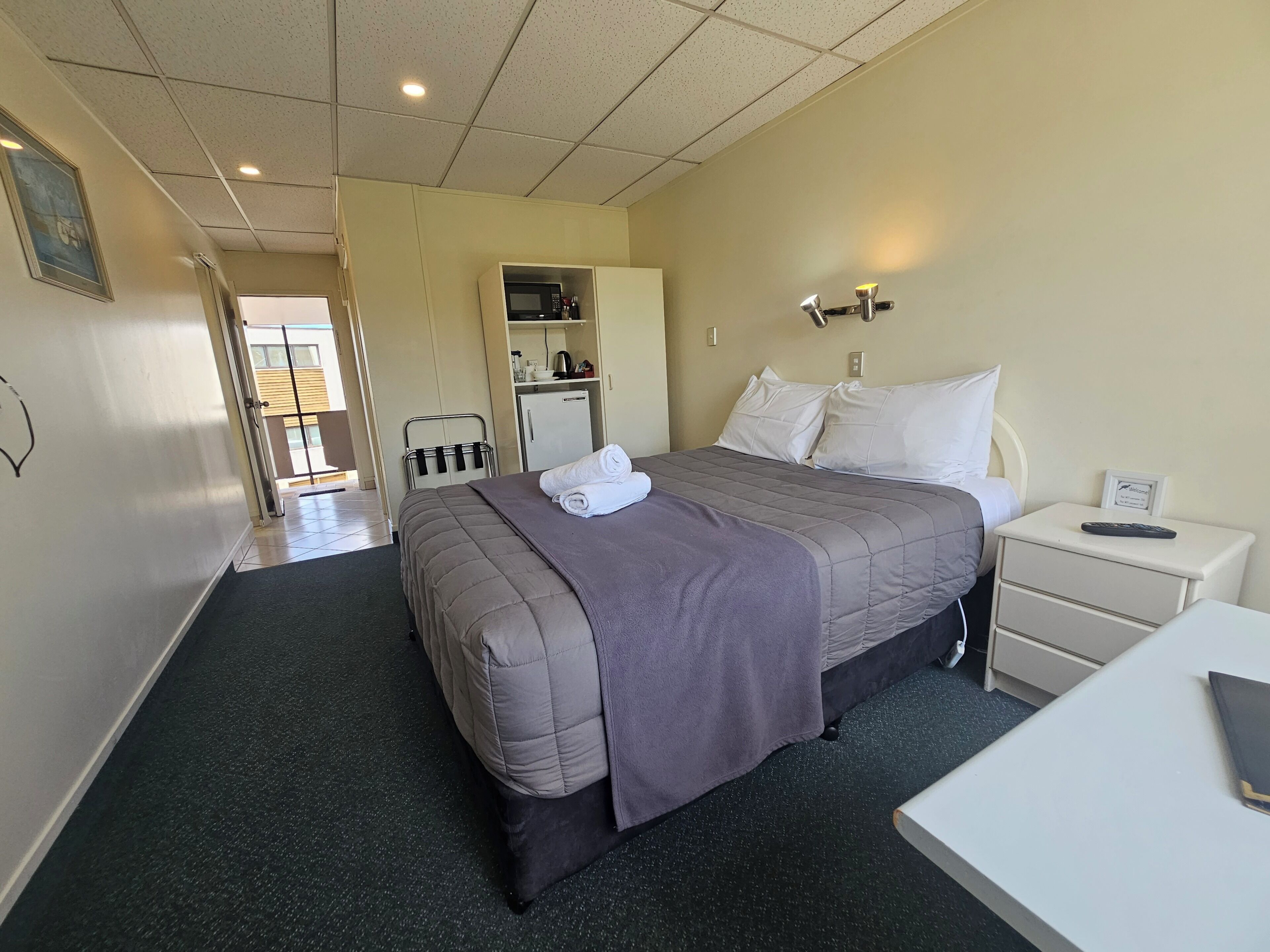 A hotel room with a bed, a nightstand with a remote, a desk, a wardrobe, and a kitchenette.