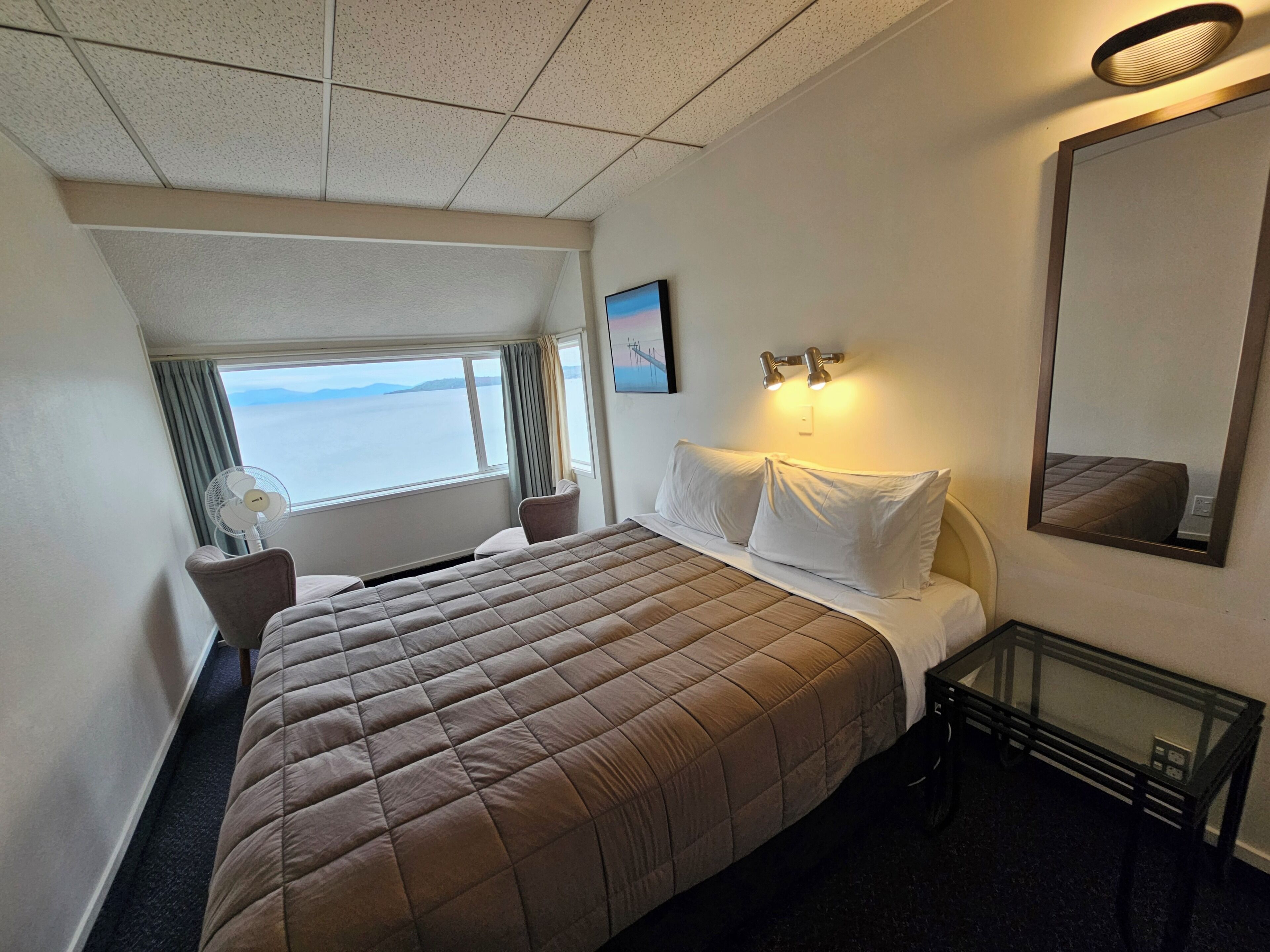 A hotel room with a bed, a bedside table, a window with curtains, a wall-mounted light, and a large mirror.