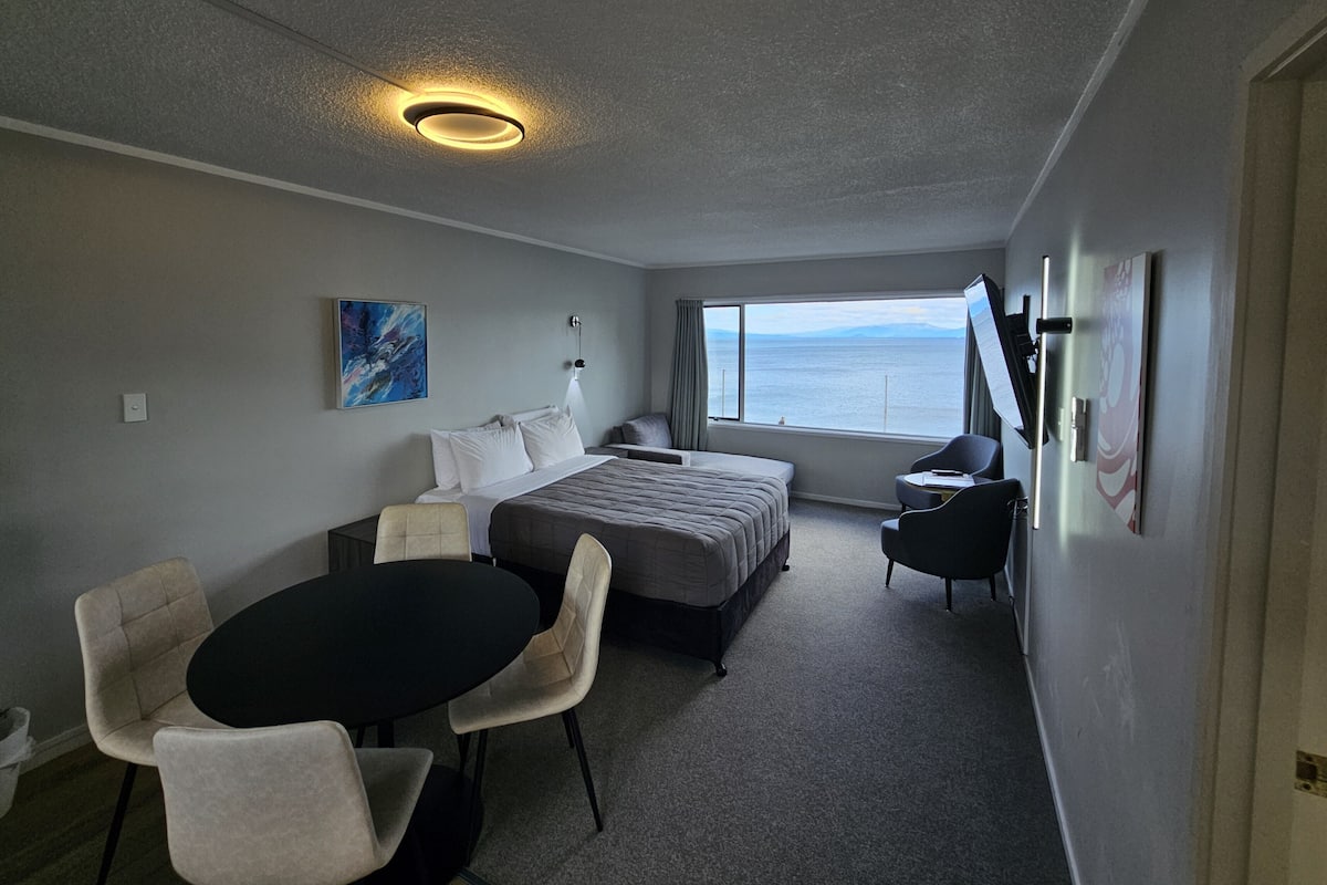 A hotel room with a bed, a desk, a chair, a table, a TV, and a window with a view of the sea.