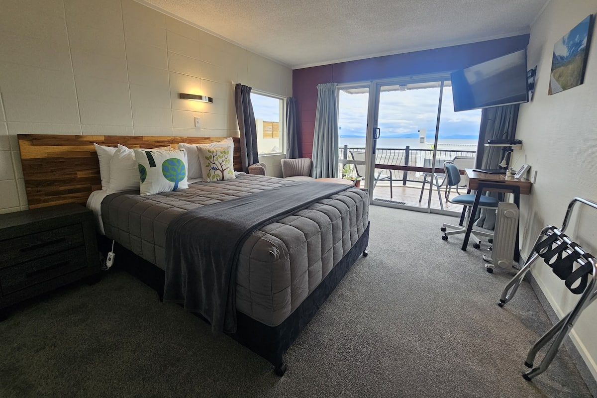A hotel room with a large bed, a desk with a chair, a TV, and a balcony with a view.