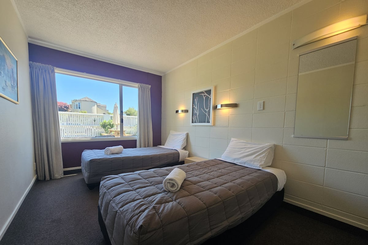 A hotel room with two beds, a large window with curtains, a view of the outside, and a wall-mounted light fixture.