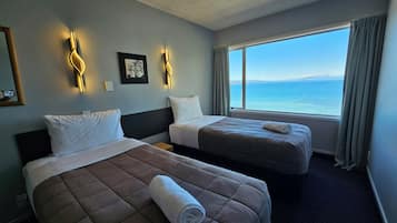 A hotel room with two beds, a large window with a view of the sea, and wall-mounted lighting.