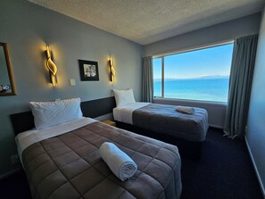 A hotel room with two beds, a large window with a view of the sea, and wall-mounted lighting. - Tui Oaks Motel (Taupo)