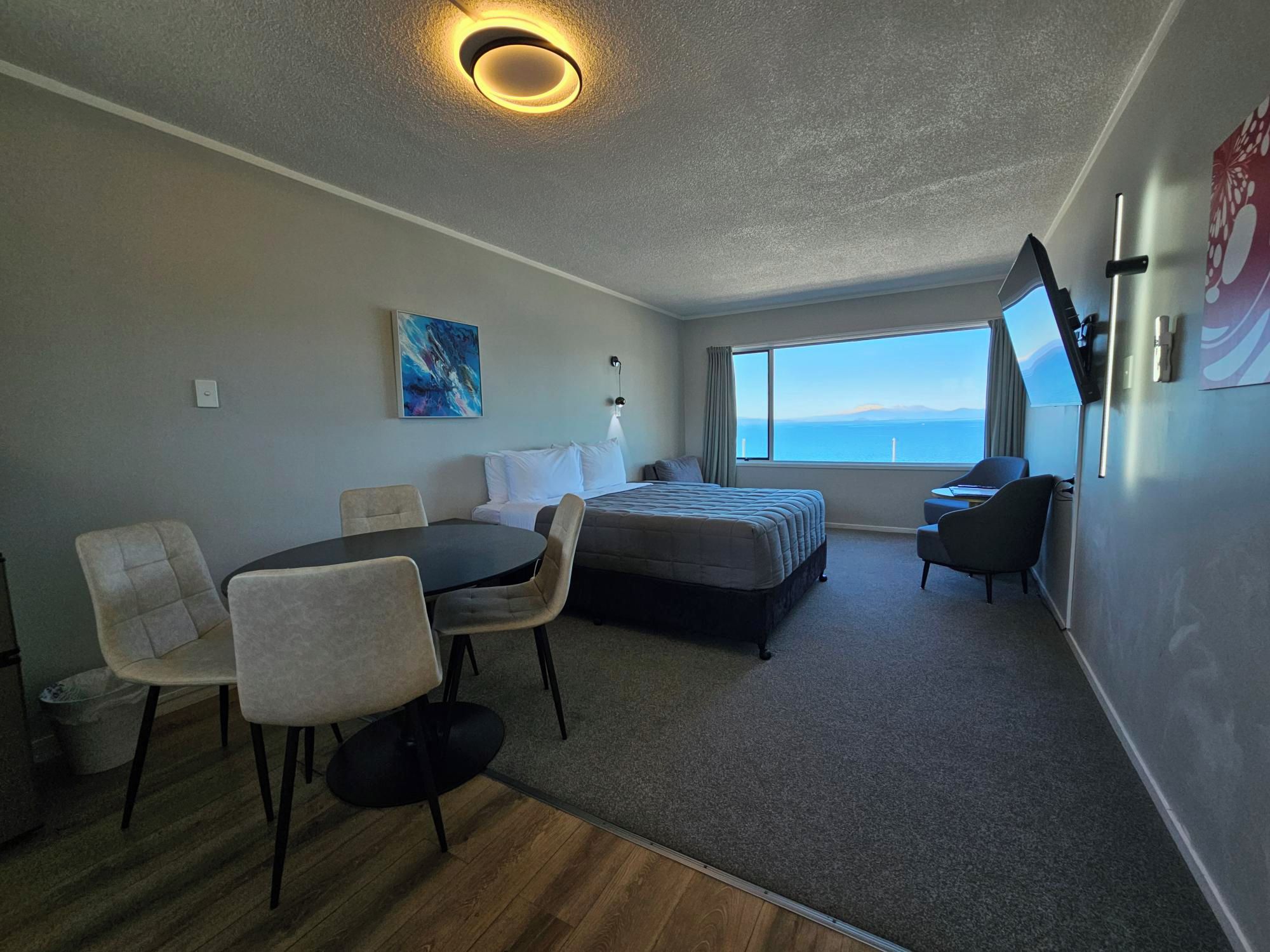 A hotel room with a bed, a dining table with chairs, a small desk, a TV, and a large window with a view.