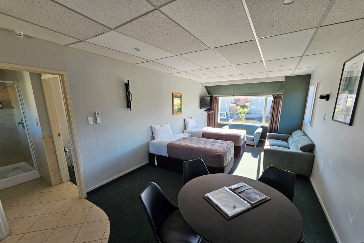 A hotel room with two beds, a sofa, a dining table with chairs, a TV, and a window with a view.