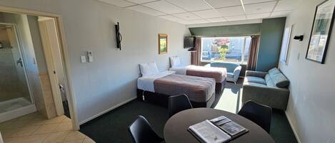 A hotel room with two beds, a sofa, a dining table with chairs, a TV, and a window with a view.