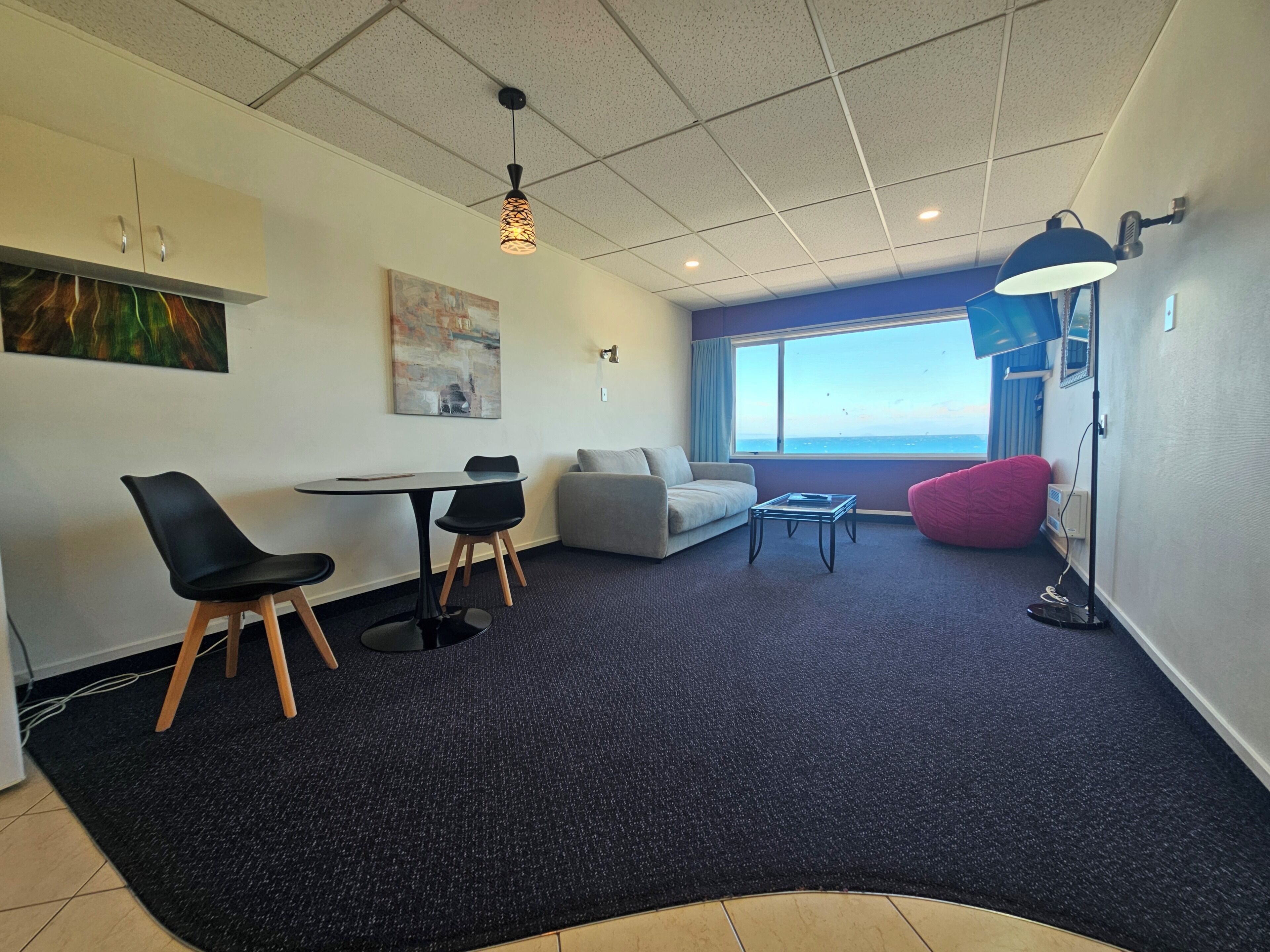 A modern living room with a dining area, a sofa, a red bean bag, a table with chairs, a lamp, and a large window with a view.