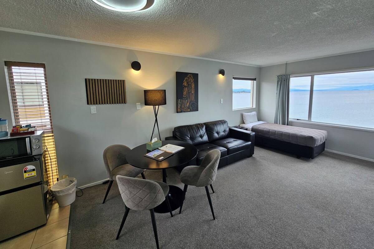 A modern living room with a sofa, a dining table with chairs, a lamp, and a view of the sea through large windows.