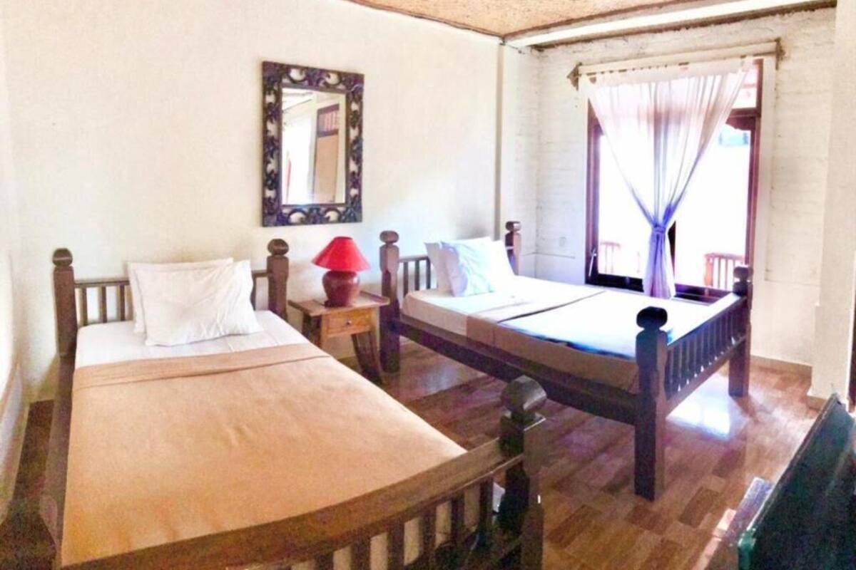 A room with two wooden beds, a nightstand with a lamp, a mirror, and a window with curtains.