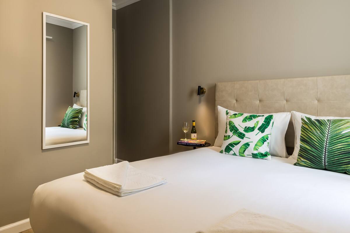 A neatly made bed with a folded white towel, a bedside table with a bottle and a glass, and a mirror on the wall.