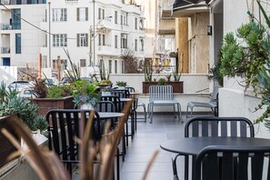 Un café al aire libre con varias mesas y sillas, rodeado de plantas en macetas y vistas a un paisaje urbano con edificios.