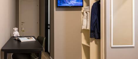 A hotel room with a TV mounted on the wall, a table with a chair, a coat rack, and a wardrobe.