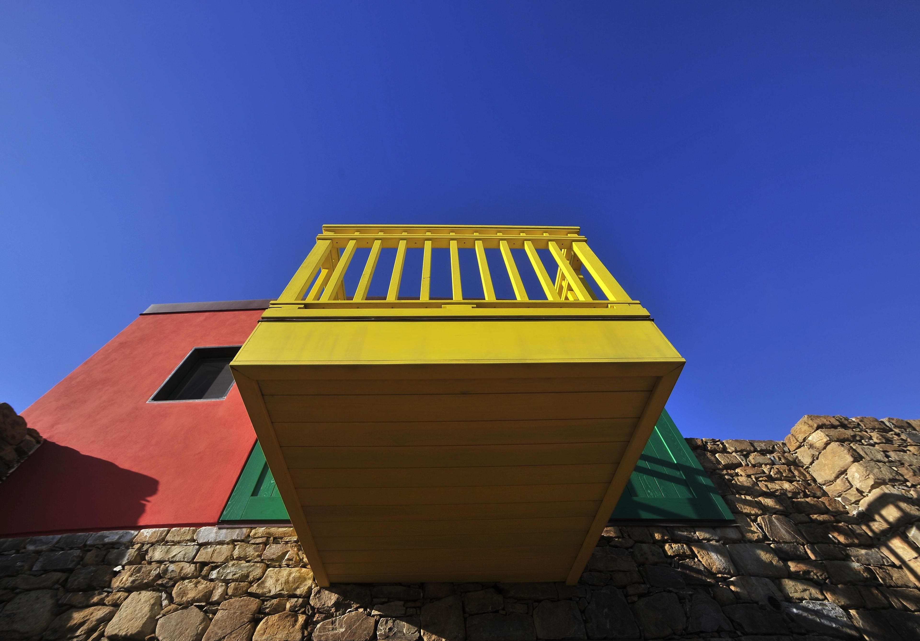 Una estructura amarilla con techo de listones, frente a un edificio rojo y un muro de piedra.