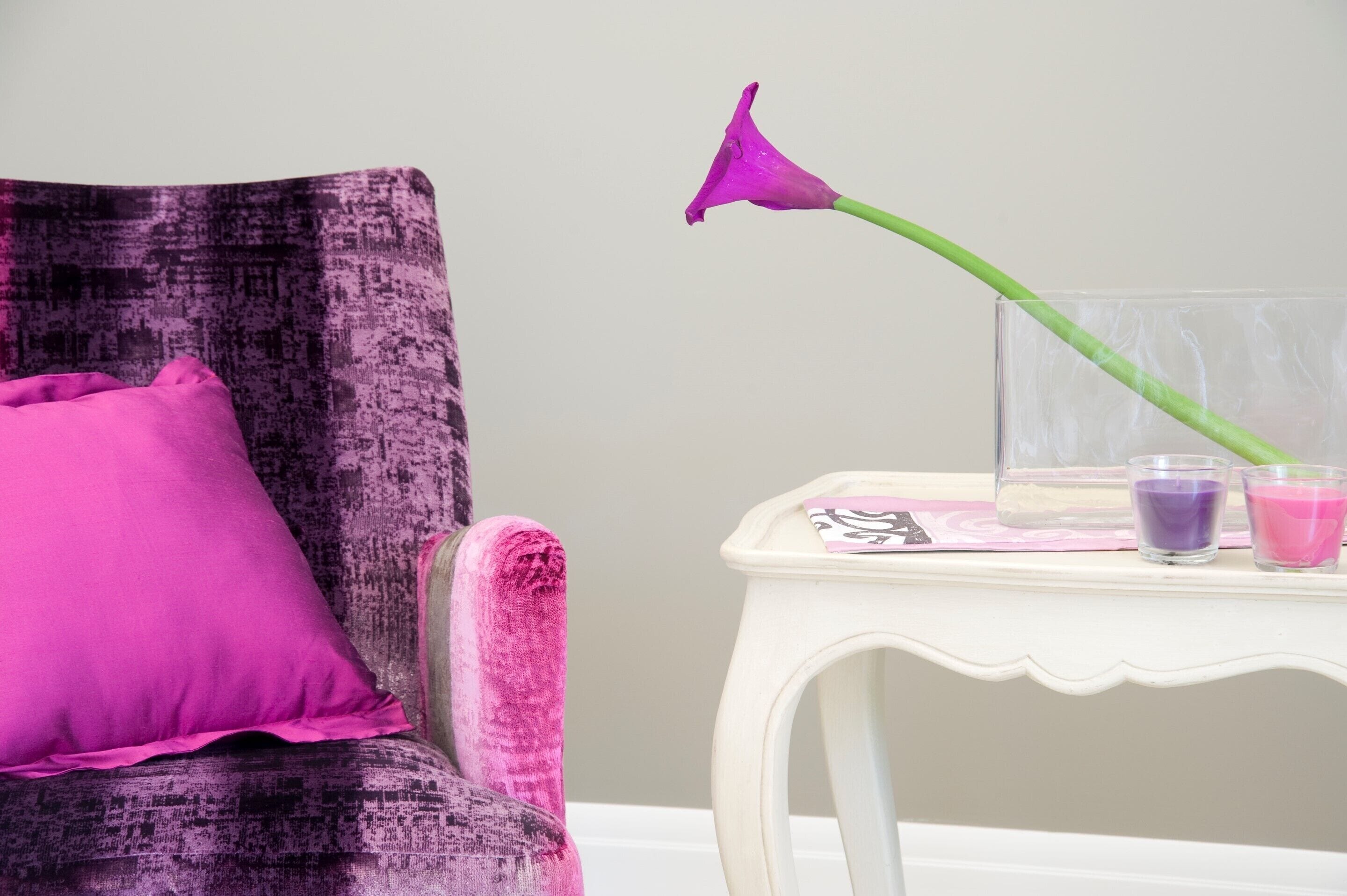 Una silla con un cojín morado y un florero de vidrio con una flor púrpura. También hay dos pequeños recipientes de vidrio sobre una mesa blanca al lado de la silla.