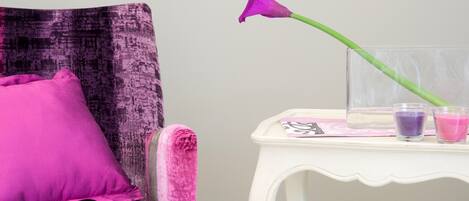 Una silla con un cojín morado y un florero de vidrio con una flor púrpura. También hay dos pequeños recipientes de vidrio sobre una mesa blanca al lado de la silla.