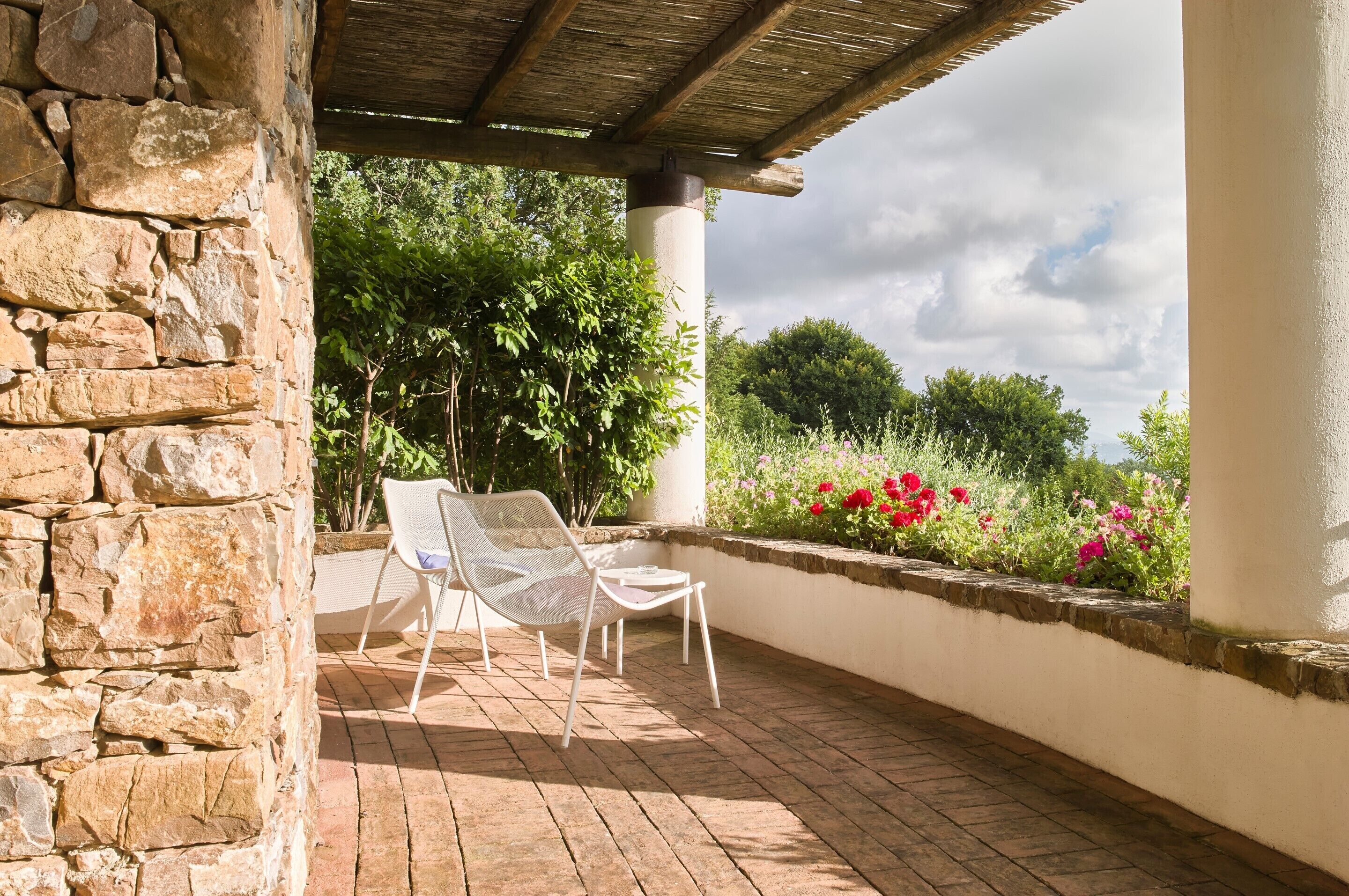 Un muro de piedra, una silla blanca y una mesa bajo una pérgola de madera en un patio con suelo de ladrillo y un parterre con flores rosas.