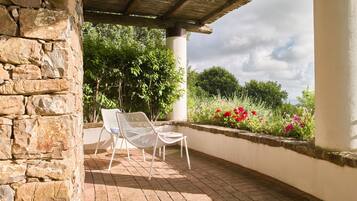 Un muro de piedra, una silla blanca y una mesa bajo una pérgola de madera en un patio con suelo de ladrillo y un parterre con flores rosas.