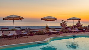 Un espace aménagé au bord de la piscine, avec des chaises longues et des parasols, offrant une vue sur une étendue d’eau au coucher du soleil.