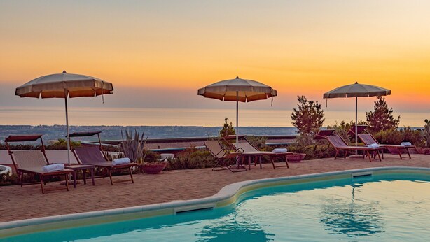 Un espace aménagé au bord de la piscine, avec des chaises longues et des parasols, offrant une vue sur une étendue d’eau au coucher du soleil.