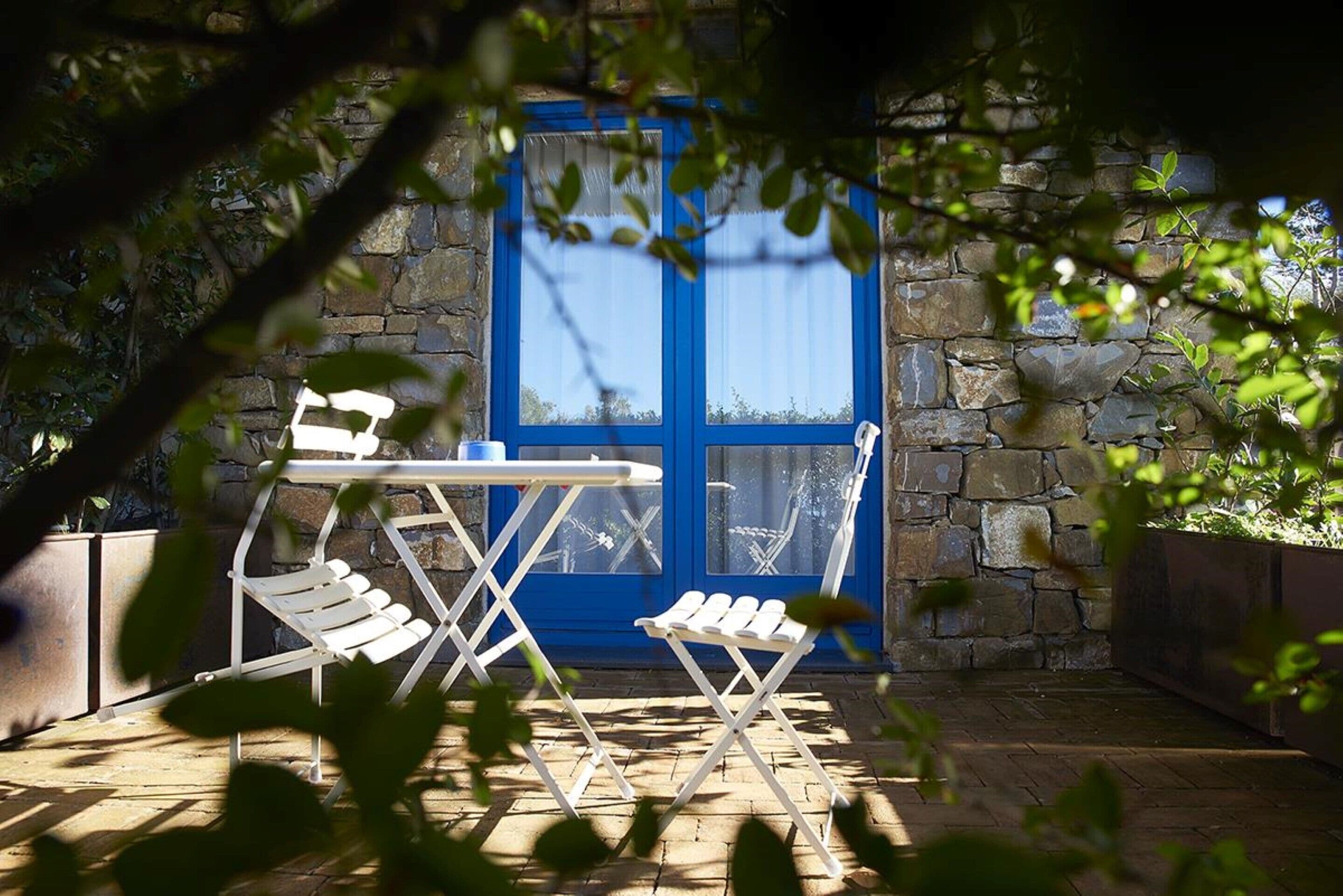 Une terrasse avec un mur en pierre, une porte bleue et des chaises pliantes blanches.