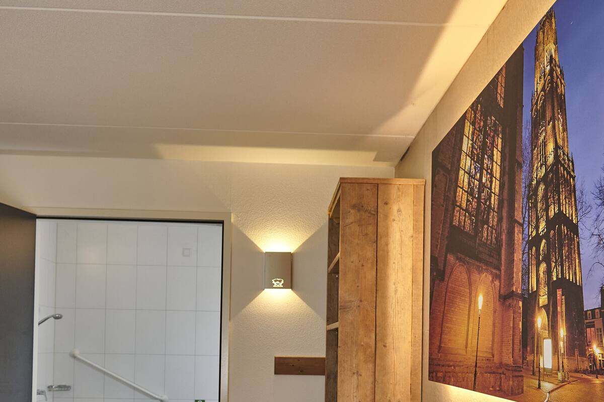 Ein Hotelzimmer mit einem Bett, einem Holzschrank, einer Wandleuchte und einem großen Fenster mit Blick auf ein Gebäude bei Nacht.