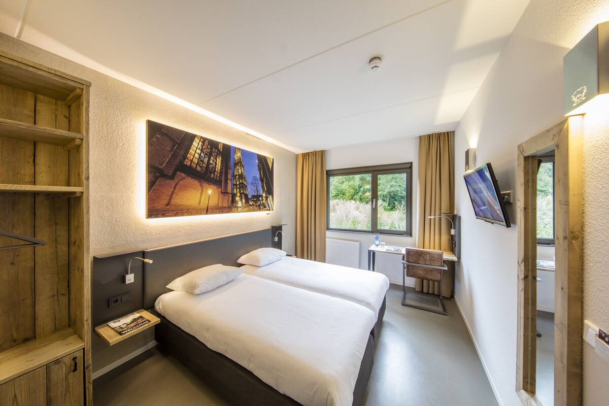 Ein Hotelzimmer mit einem großen Bett, einem Schreibtisch, einem Fernseher und einem Fenster mit Vorhängen.