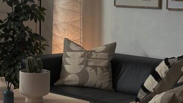 Een woonkamer met een donkere bank, een ronde tafel, een lamp, een cactus en een poster aan de muur.