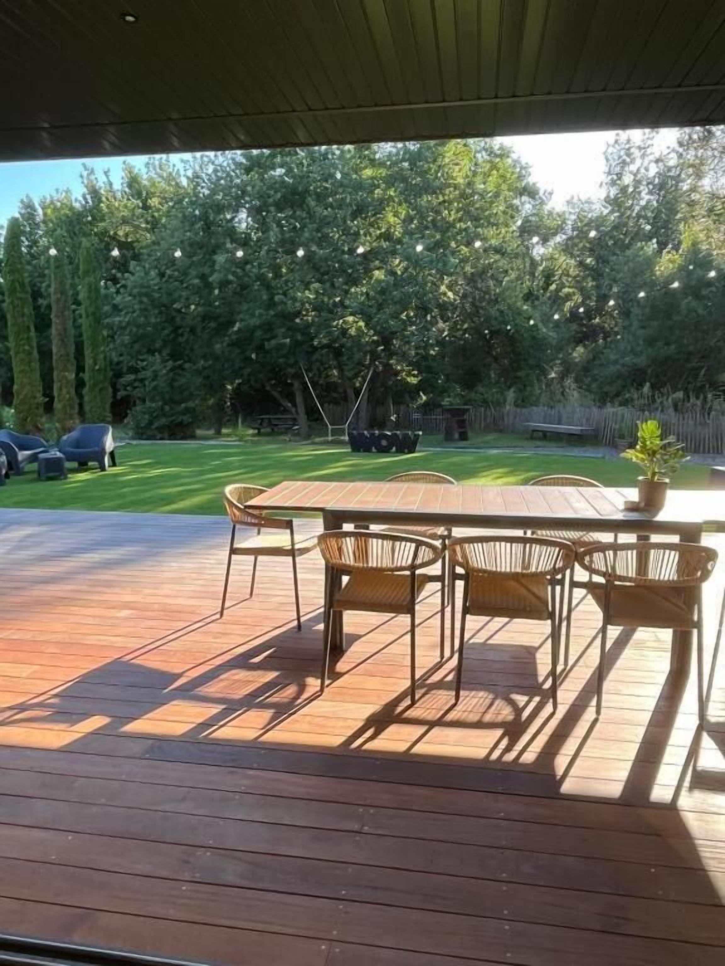 Una terraza de madera con una mesa y sillas, rodeada por un césped y árboles.