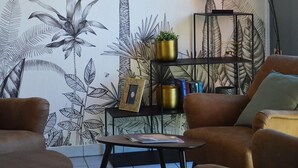 Ein Wohnzimmer mit einer großen Wandmalerei von tropischem Grünwerk, einer modernen Lampe, einem Bücherregal und zwei Sofas.