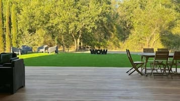 Une terrasse extérieure avec une plateforme en bois, un espace salon comprenant des chaises et une table, et une vue sur une pelouse verdoyante et des arbres.