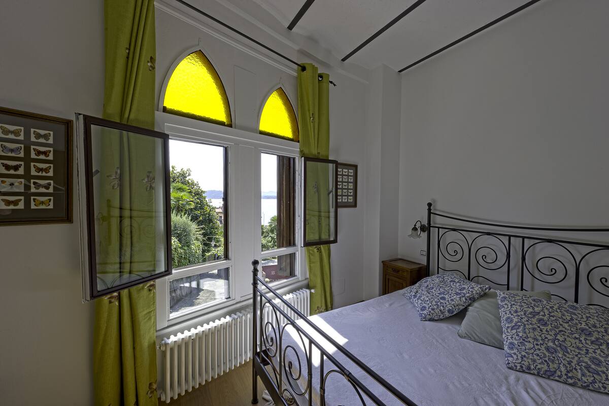Un dormitorio con una cama, dos ventanas con cortinas verdes, un radiador y cuadros de mariposas en la pared.