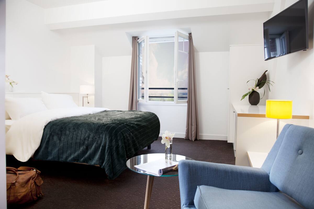 A hotel room with a bed, a chair, a small table, a TV, a window with curtains, and a desk with a lamp.