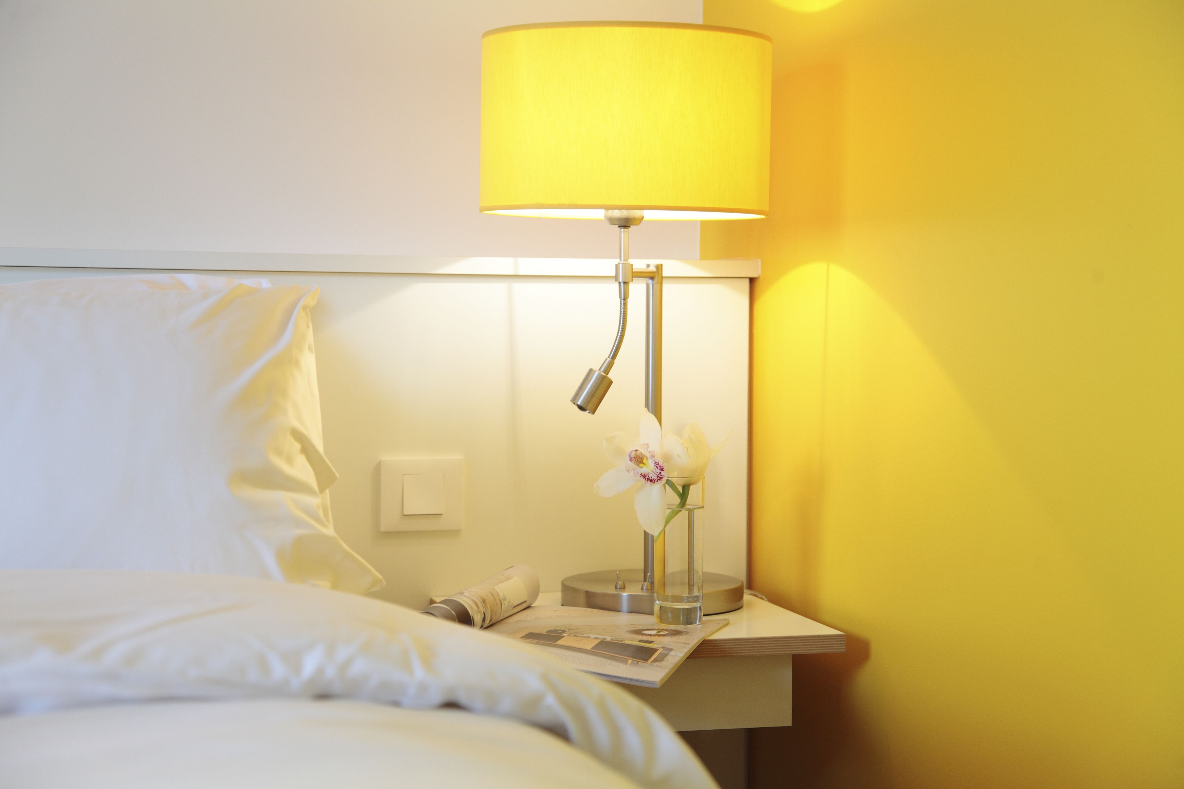 Quarto de hotel com uma cama, mesa de cabeceira com abajur, um vaso com uma flor e uma revista.