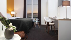 Habitación de hotel con cama, escritorio, silla, una mesita con una botella y un vaso, y vista al exterior a través de una ventana.