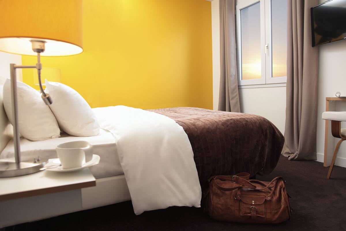 A hotel room with a bed, a bedside table with a lamp, a cup, and a brown bag.