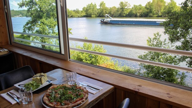 Una mesa de comedor con pizza y ensalada, una ventana con vistas al río y a un barco, y una barandilla de madera.