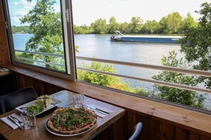 Una mesa de comedor con pizza y ensalada, una ventana con vistas al río y a un barco, y una barandilla de madera.