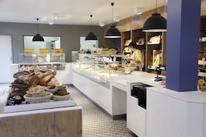 A bakery with a variety of baked goods displayed in glass cases and on shelves, a counter with a person working, and pendant lighting.